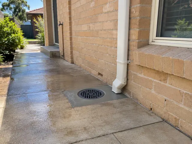 Outdoor drain grate set in concrete paving beside a brick house wall in Waitara
