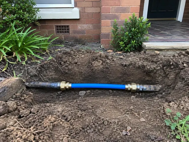 Exposed repaired water pipe in a dirt trench beside a brick house exterior in Asquith